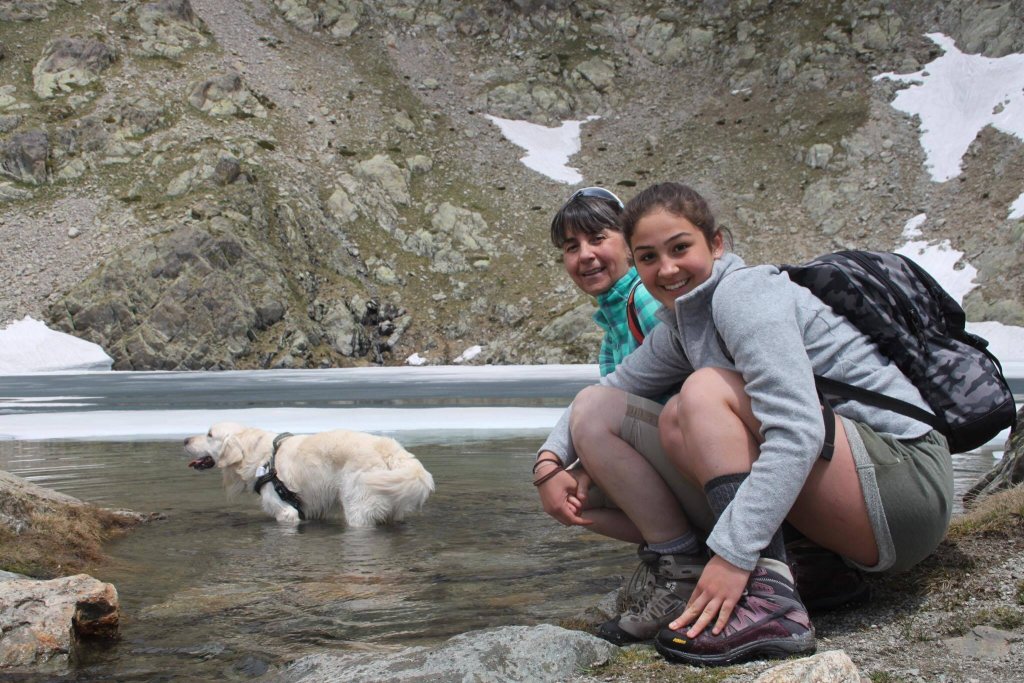 Two women hiking kneeling down next to golden retriever dog in a lake in the mountains 