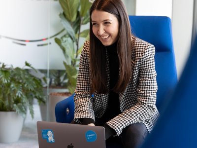 woman with brown hair sitting in a blue chair with laptop laughing