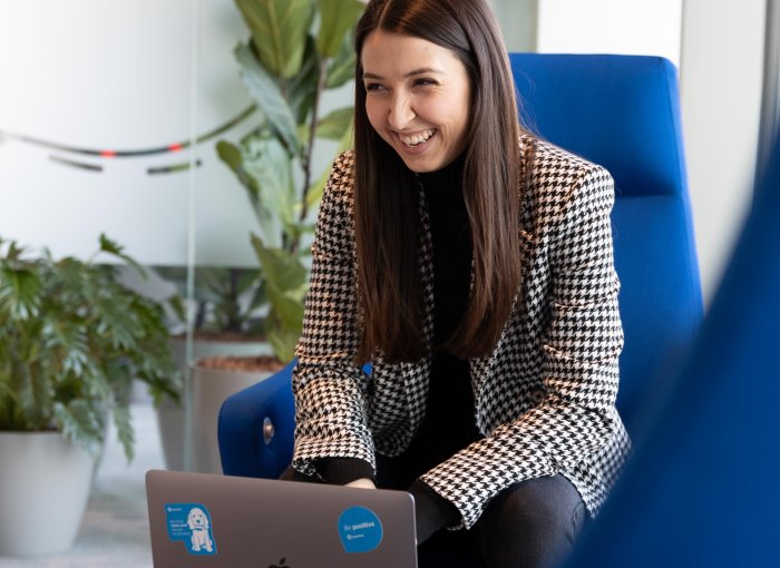 woman with brown hair sitting in a blue chair with laptop laughing