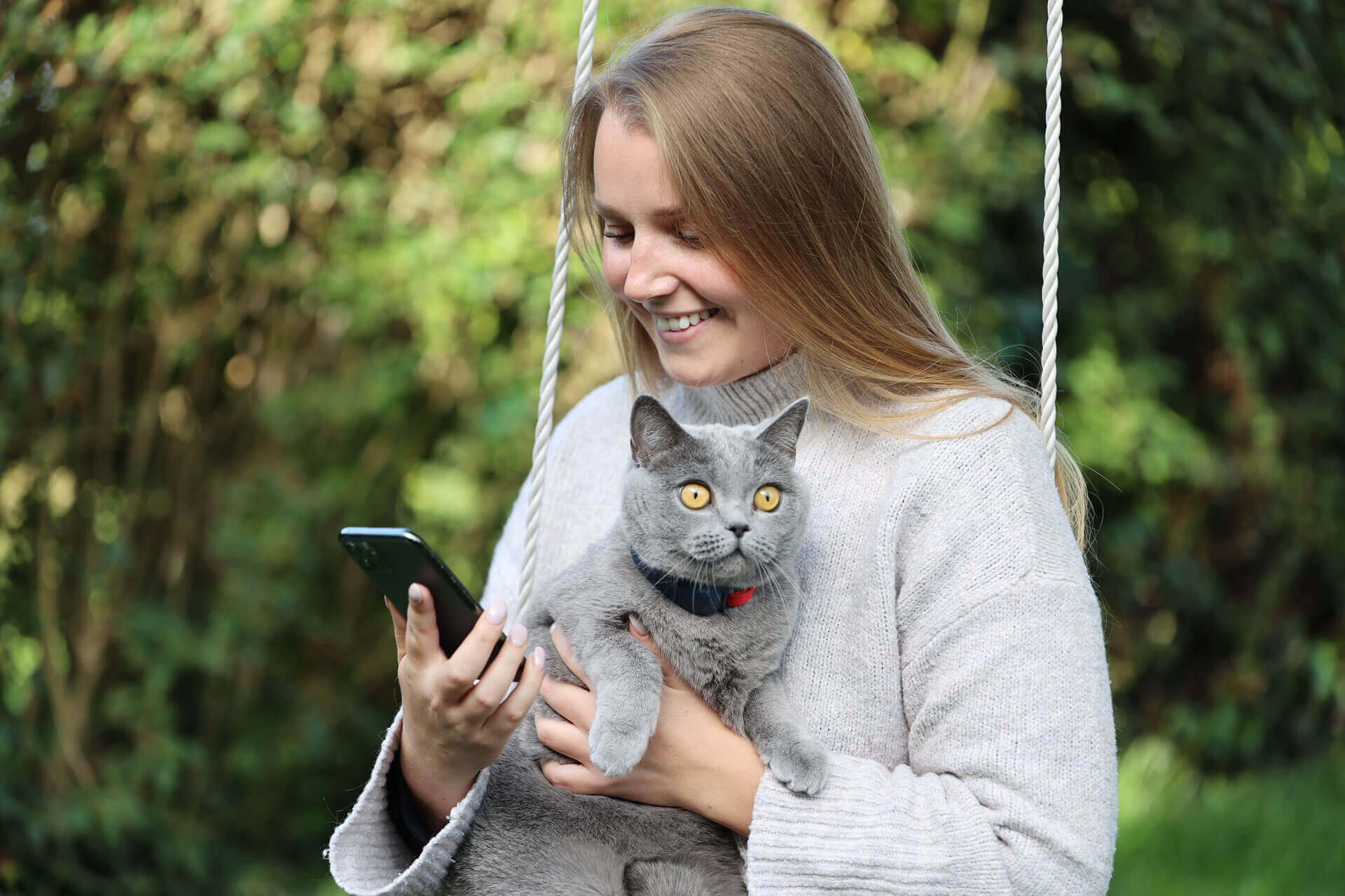 Nainen sylissään harmaa kissa, jolla on GPS-kissapaikannin