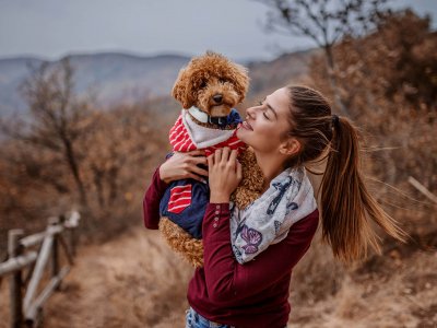 femme souriante debout dans la nature serrant un petit chien avec foulard dans ses bras