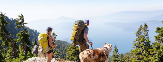 Zwei Wanderer mit Rucksack und Hund stehen auf einem Bergfelsen und genießen das Panorama
