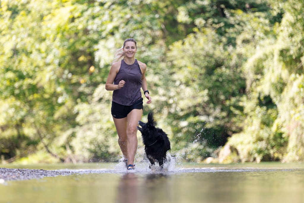 Läuferin joggt mit ihrem Hund durch seichtes Wasser im Wald