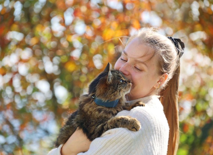 en kvinne holder en brun katt iført gps-tracker for katt, ute med et tre i bakgrunnen
