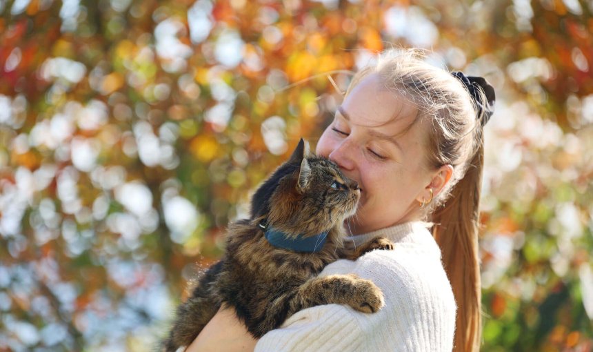 Mujer abrazando un gato marrón con un localizador gps en el collar al aire libre, con un árbol en el fondo