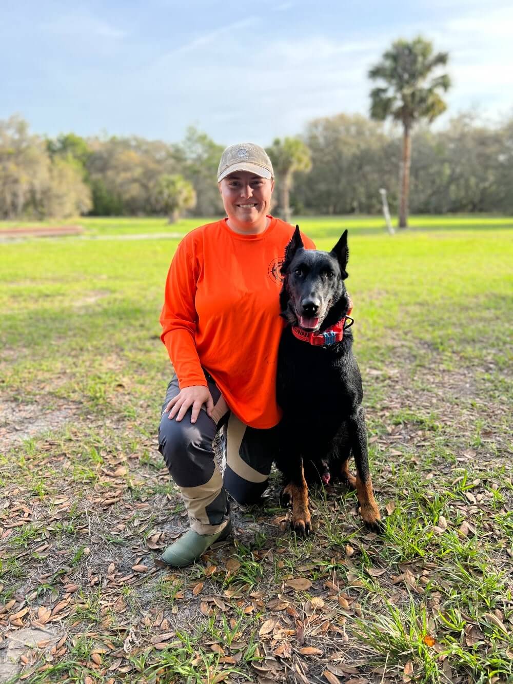 woman in orange shirt kneeling next to her K9 SAR dog wearing a GPS tracker outside