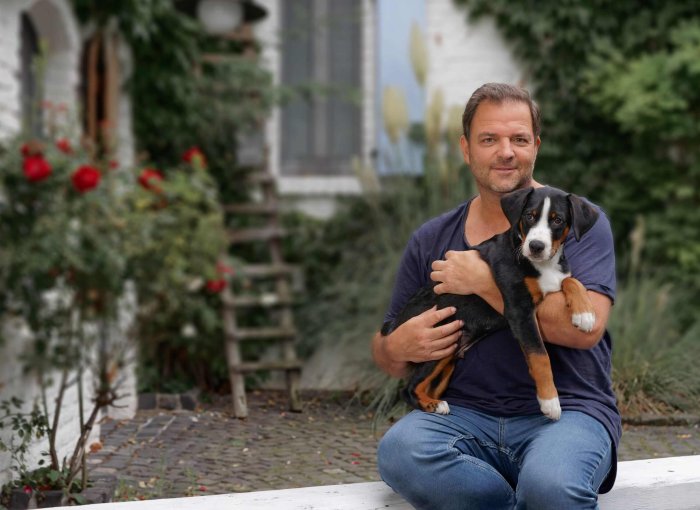 Martin Rütter sitzt vor einem Haus mit einem Welpen auf dem Schoß
