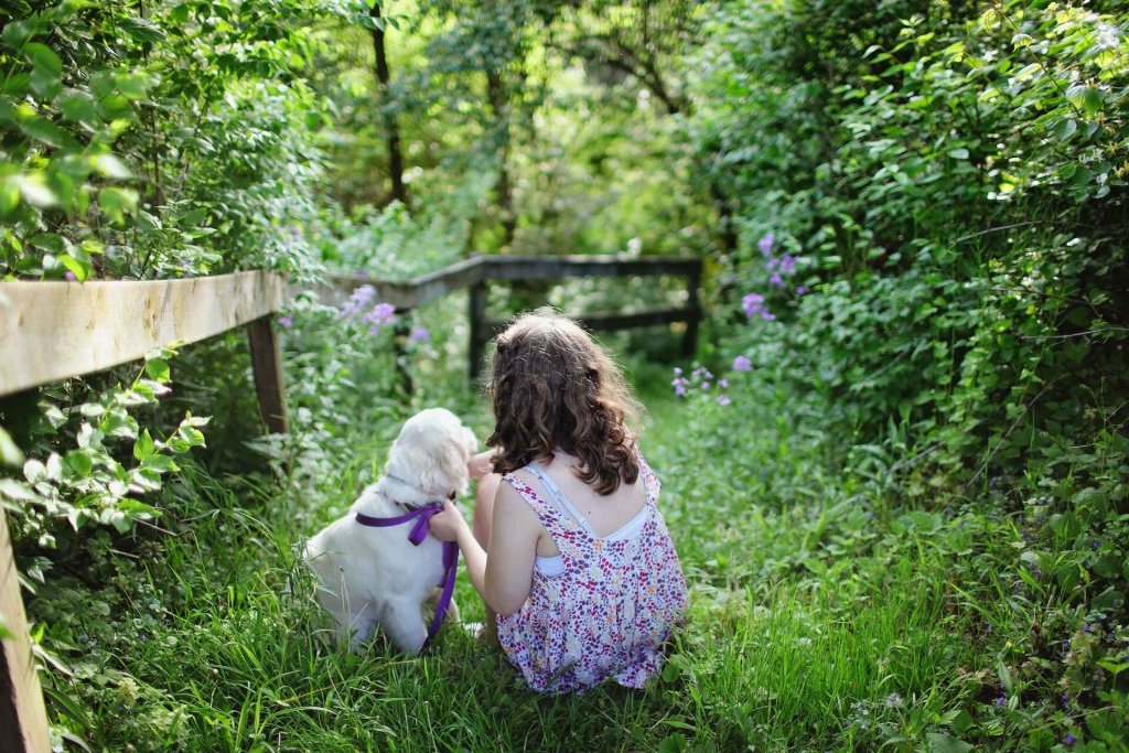 A puppy and a girl sit together in a garden surrounded by leafy plants