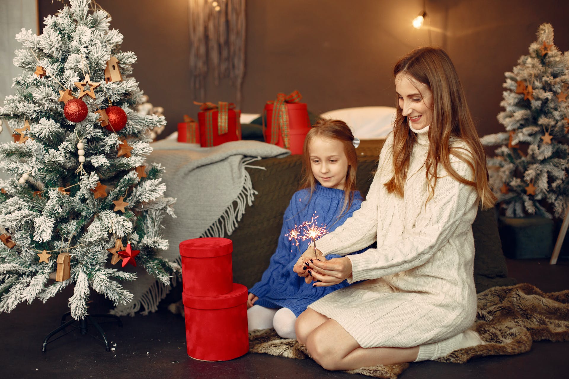 A woman and girl lighting a Christmas sparkler by a tree indoors