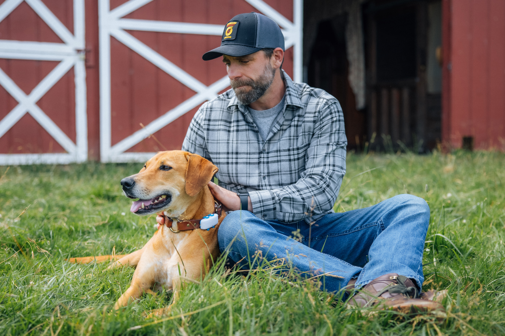 dog owner sitting in front of a barn in the grass with his dog with a tractive gps tracker on his collar