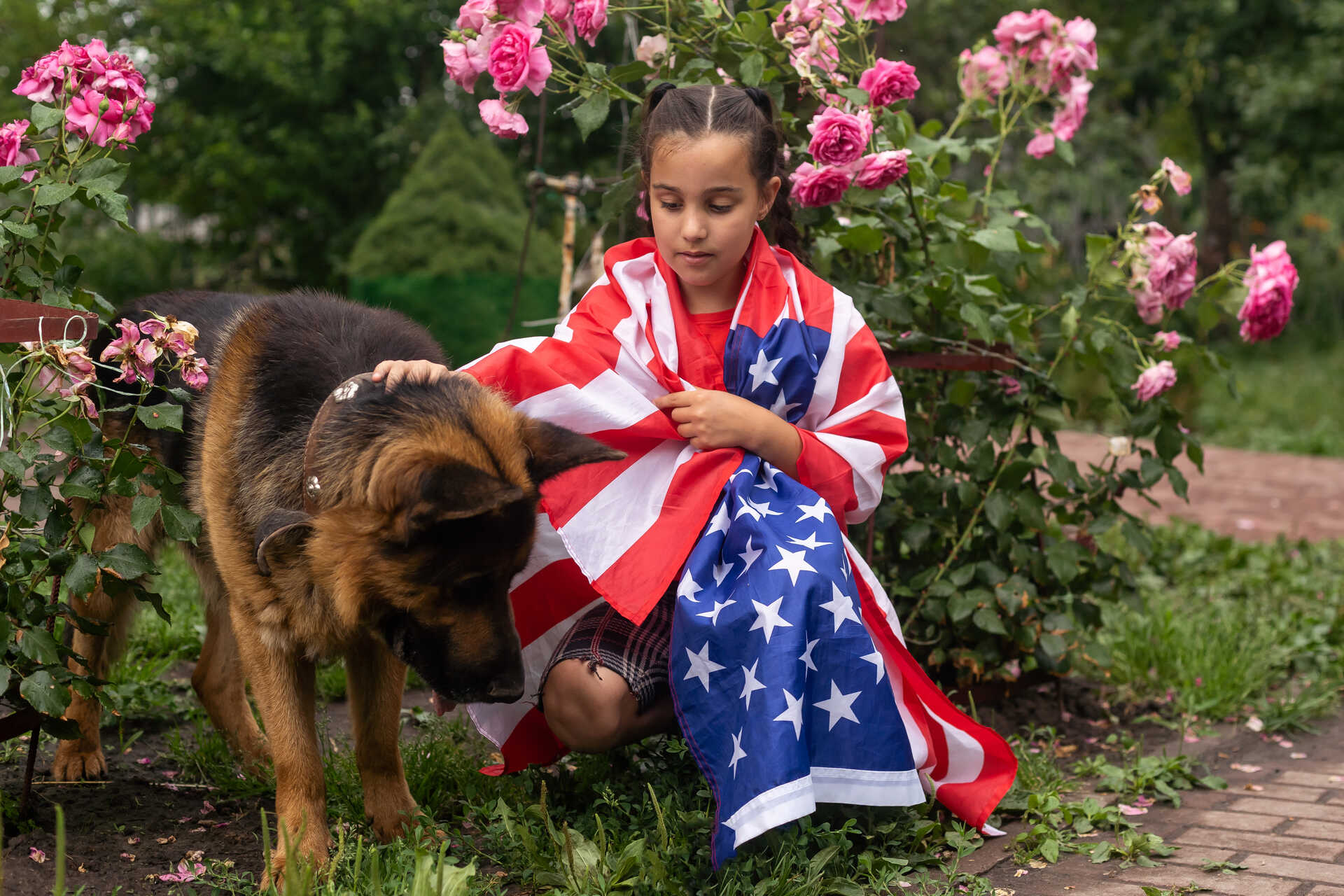 A girl wrapped in a US flag sitting by a dog in a garden